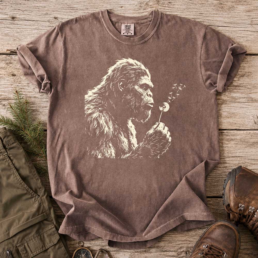 May include a Espresso Bigfoot blowing a dandelion graphic on a casual T-shirt for men and women with a funny Sasquatch blowing wishes design.