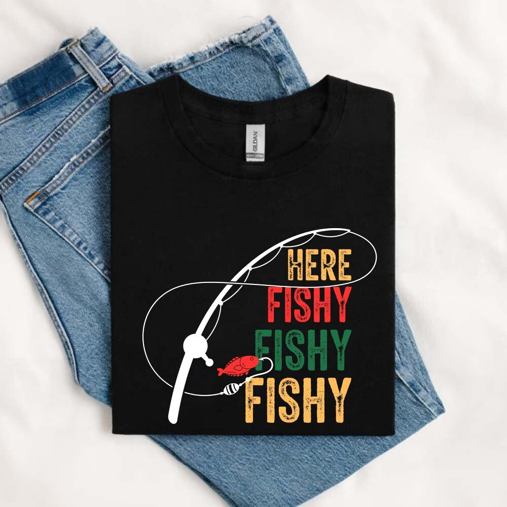 May include an image of a folded black unisex T-shirt with a colorful “HERE FISHY FISHY FISHY” text design and fishing rod graphic, styled beside denim jeans for a casual fisherman look.