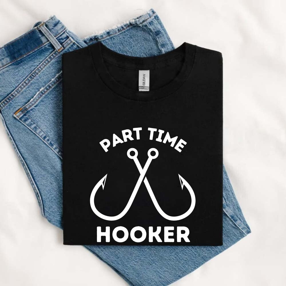 May include an image of a folded black unisex T-shirt with two crossed fishing hooks and the text “PART TIME HOOKER” printed in bold white lettering, styled next to denim jeans for a casual fishing-inspired outfit.